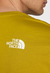Majica kratkih rukava, okruglog izreza u boji gorušice, izrađena od teksturiranog materijala. Ima bijeli logo "THE NORTH FACE" na gornjem dijelu leđa.