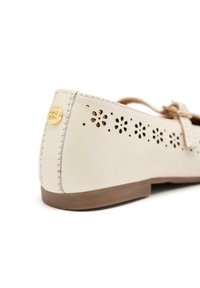 Zapato plano de cuero beige con recortes florales a lo largo del costado, suela marrón y detalles de costura decorativos. Presenta un pequeño logo dorado.
