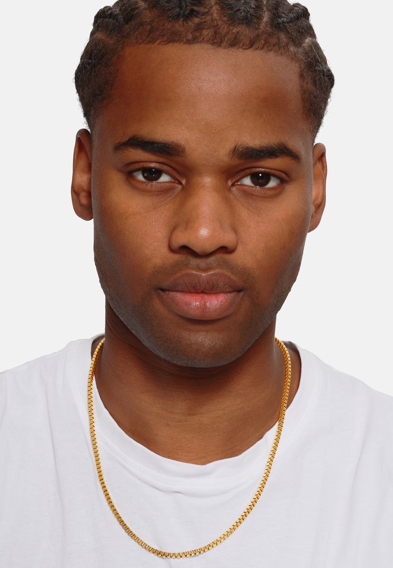 Collier en or avec un design plat et entrelacé, reposant sur un T-shirt blanc uni, contraste avec une texture de peau lisse.
