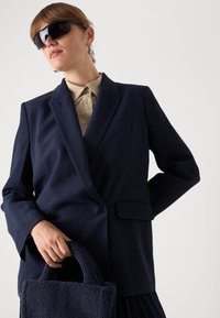 Námořnický blazer s proužky, texturovaná námořnická koženková kabelka, béžová košile, černé sluneční brýle. Blazer má klopové límce a klopové kapsy.