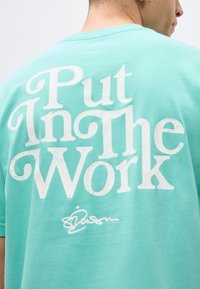 Άτομο που φοράει τιρκουάζ πουκάμισο με μεγάλο λευκό κείμενο στην πλάτη που γράφει "Put In The Work" και μια μικρή υπογραφή από κάτω.