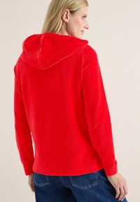 Femme portant une veste à capuche rouge vif avec des manches longues, vue de dos, associée à un jean en denim bleu.