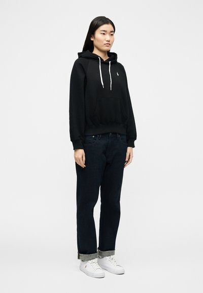 Γυναίκα που στέκεται φορώντας μαύρο φούτερ με λευκά κορδόνια, σκούρο τζιν με γυρισμένα μανίκια και λευκά αθλητικά παπούτσια, μπροστά σε απλό λευκό φόντο.