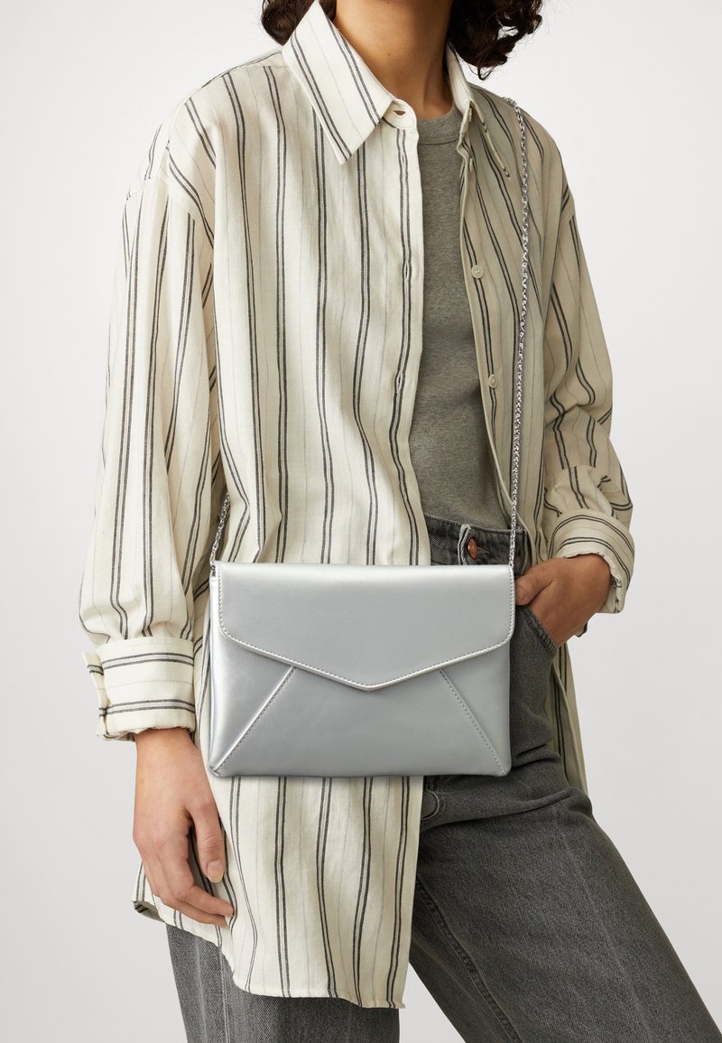 Silberne metallische Umschlag-Crossbody-Tasche mit einer Kettenriemen. Glatte Textur und gestickte Akzente. Kombiniert mit einem gestreiften Shirt und grauen Jeans.