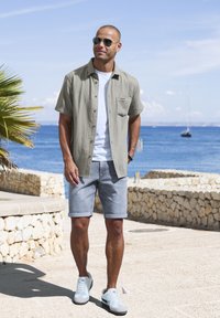 Camicia verde chiaro a maniche corte con taschino, t-shirt bianca sotto, pantaloni corti in denim grigio e sneakers grigie/bianche in un ambiente costiero.