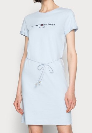 Kobieta w jasnoniebieskiej sukience z krótkim rękawem z logo "Tommy Hilfiger EST. 1985" i paskiem w talii ozdobionym drobnymi złotymi koralikami.