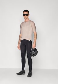 Maillot de cyclisme beige à manches courtes, collants rembourrés noirs, chaussures noires, tenant un casque noir, tatouages visibles sur le bras, design minimaliste.