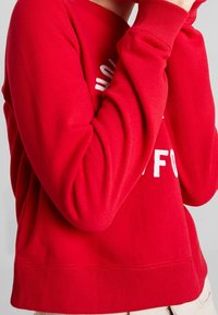 Sudadera de algodón roja con dobladillo y puños acanalados. Presenta texto en blanco en la parte delantera. Textura suave, diseño casual y corte ligeramente ajustado.