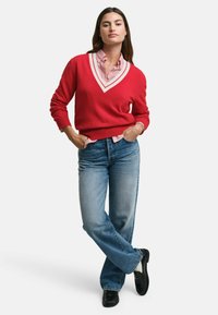 Roter V-Ausschnitt-Pullover mit weißen Kanten, darüber getragen ein rot-weiß gestreiftes Hemd. Kombiniert mit hellblauen Jeans und schwarzen Schuhen.