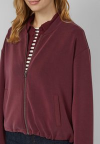 Frau in einer bordeauxfarbenen Zip-Jacke über einem gestreiften weinroten und weißen Shirt mit dunkelblauen Jeans vor einem schlichten grauen Hintergrund.