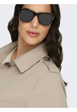 Kvinde iført briller med skildpaddemønster og beige trenchcoat med knaplukket skulderklaffe, neutral baggrund.