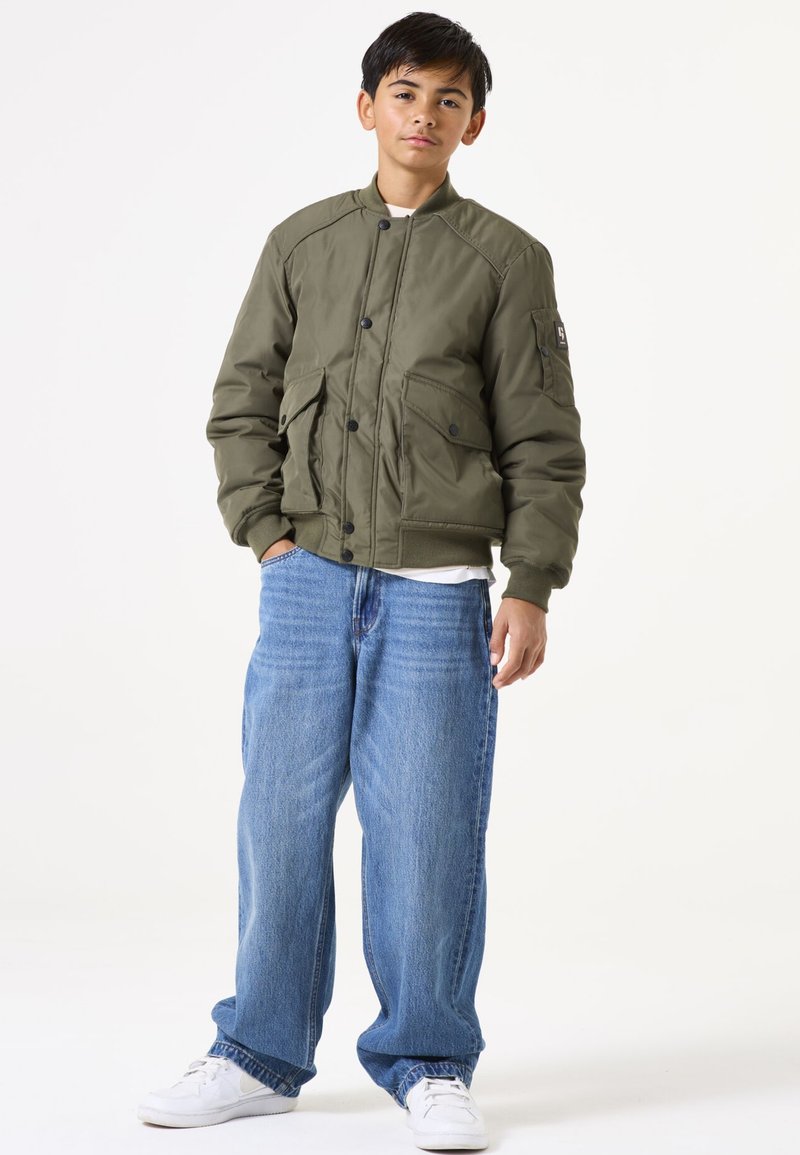 Olivgrüne Bomberjacke mit Reißverschluss, Druckknöpfen und zwei vorderen Taschen; kombiniert mit locker sitzenden blauen Jeans und weißen Turnschuhen.