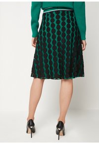 Falda verde con lunares negros, hasta la rodilla, combinada con un suéter verde y zapatos de tacón negro, vista desde atrás.