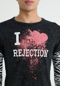Černá košile s dlouhým rukávem, která má růžové srdce ve stylu rozstřiku a bílo-černý nápis "I REJECTION." Rukávy s černo-bílým zebrovým vzorem. Materiál: bavlna.
