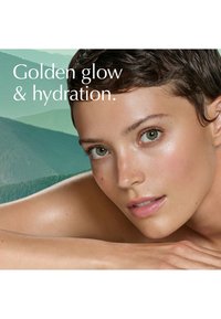 Femme aux cheveux courts et foncés reposant sa peau éclatante et hydratée contre son bras, avec un doux paysage de montagnes vertes en arrière-plan.