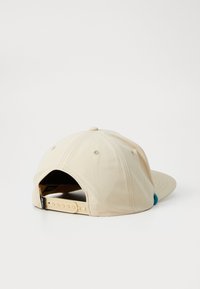 Beige cap med buet skygge, justerbar stropp bak og turkis aksent. Laget av stoff med synlige sømdetaljer.