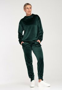 Zestaw loungewear w zielonym aksamicie z dekoltem w formie drapowania, długimi rękawami i elastycznymi mankietami, w połączeniu z dopasowanymi spodniami i białymi sneakersami.