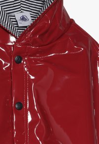 Czerwona, laminowana kurtka przeciwdeszczowa o błyszczącej powierzchni, z czarnymi guzikami i kontrastową, paskowaną podszewką.