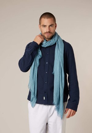 Homme portant une chemise bleu foncé boutonnée, un pantalon blanc et une écharpe bleu clair, ajustant son écharpe d'une main sur un fond uni.