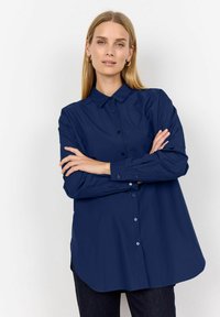 Tamsiai mėlynas ilgomis rankovėmis mygtukais užsegamas marškinis su smailiu apykakle, tiesiu apatine dalimi ir laisvu siluetu. Lygus, lengvas audinys.