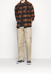 Braun-und-schwarz kariertes Flanellhemd, beigetöne Hose mit Knieflicken und schwarze Schuhe mit weißen Akzenten. Lässiges Outfit mit einem gestuften Design.