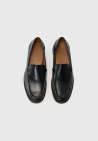 Mocasines de cuero negro con un acabado suave, punta redondeada, costuras visibles y un collar acolchado. El interior presenta un forro en tono marrón claro.