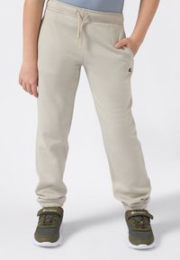 Kind trägt beigefarbene Sweatpants mit Kordelzug und Champion-Logo, kombiniert mit grünen und weißen Champion-Sneakers, steht mit entspannten Händen.