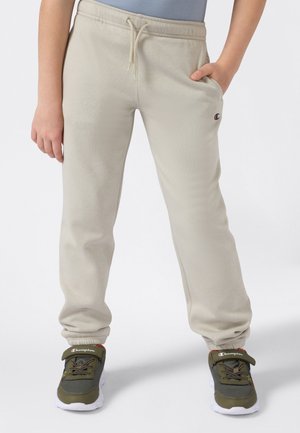 Barn iført beige joggebukser med snor og Champion-logo, kombinert med grønne og hvite Champion-sneakers, stående med avslappede hender.