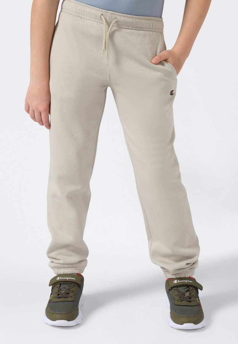Kind trägt beigefarbene Sweatpants mit Kordelzug und Champion-Logo, kombiniert mit grünen und weißen Champion-Sneakers, steht mit entspannten Händen.