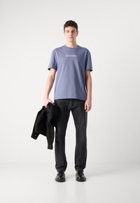 Pelēka T-kreklu ar baltu "Calvin Klein" tekstu, melniem džinsu biksēm, tur melnu jaku un valkā melnas kurpes; minimālistisks dizains.