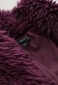 Lilla faux fur materiale med en fluffy tekstur, der viser en indvendig foret i en lysere lilla tone. Etiketten viser "NEXT" med plejeinformation.