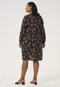 Robe noire avec imprimé floral, manches longues, taille froncée et longueur intermédiaire. Associée à des talons ouverts noirs.