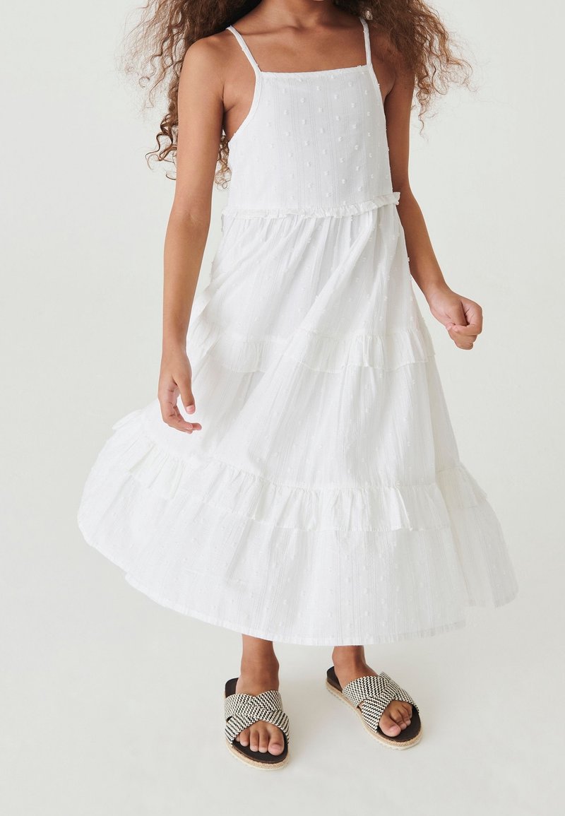 Robe maxi blanche avec des volants superposés, tissu texturé comprenant de petits points en relief, et fines bretelles ajustables. Associée à des sandales noires à motifs.