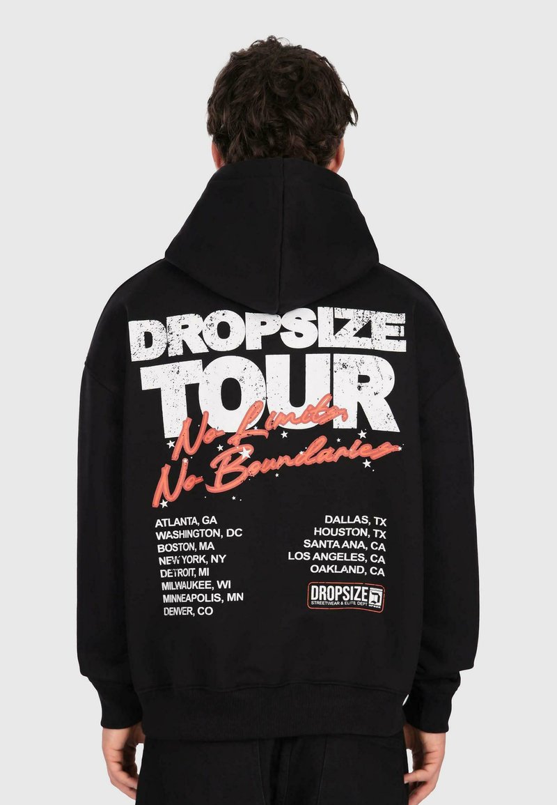 Osoba v čiernej mikine s kapucňou, na ktorej sú biele a oranžové nápisy "DROPSIZE TOUR No Limits No Boundaries" a na zadnej strane zoznam miest v USA.