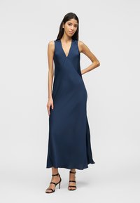 Γυναίκα με αμάνικο μάξι φόρεμα σε σκούρο μπλε απόχρωση με V-λαιμόκοψη και πλαϊνές σκισμές, φοράει μαύρες ψηλοτάκουνες πέδιλα με λουράκια, στέκεται με το ένα χέρι στη μέση.