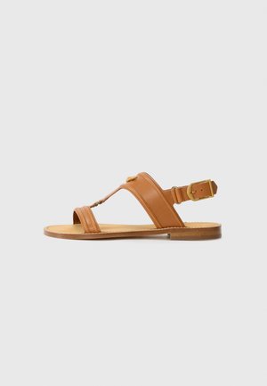 Brown leather sandals with adjustable ankle strap, flat sole, and crossed upper straps. Smooth texture, minimal design, gold hardware accent.