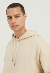 Sudadera con capucha beige hecha de tela suave con capucha ajustable con cordón, presenta un ajuste relajado y detalles sutiles de costuras en los hombros.