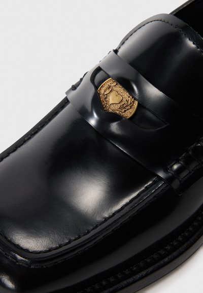 Scarpa mocassino in pelle nera con finitura lucida, emblema dorato con design decorativo e dettagli di cucitura lungo la suola.