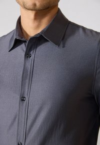 Camicia button-up grigio scuro con colletto classico, caratterizzata da una texture liscia e bottoni neri lucidi. Vestibilità su misura, design minimalista.