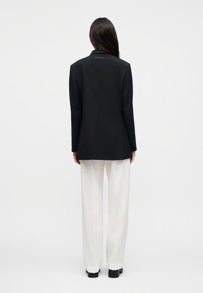 Μαύρο σακάκι σε στενή γραμμή με μακριά μανίκια, γιακά με εγκοπή και απαλό υφή, συνδυασμένο με λευκό παντελόνι φαρδιάς γραμμής και μαύρες ψηλοτάκουνες γόβες.