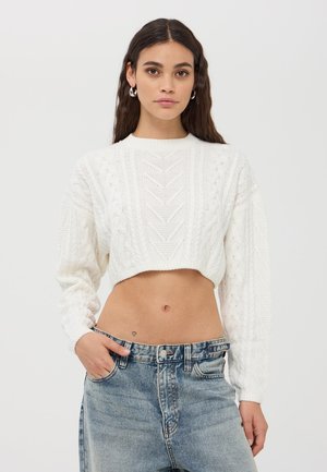 Maglione bianco corto con motivi a trecce testurizzati e maniche lunghe, abbinato a jeans di jeans sbiaditi a vita alta.