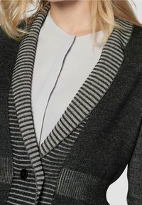 Černý svetr s pruhovanými šedými klopami a kapsami, vyrobený z texturované látky a s knoflíkovým zapínáním, vrstvený přes světle šedý top.