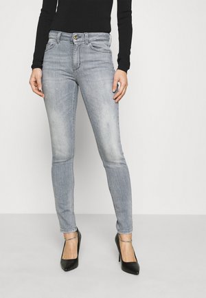 Frau trägt hellgraue Skinny-Jeans, schwarzen Langarm-Top und schwarze High Heels mit Fußkettchen, steht vor einfarbigem Hintergrund.