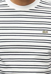 Κοντινό πλάνο λευκού t-shirt με λεπτές μαύρες οριζόντιες ρίγες και μικρό πράσινο λογότυπο κροκοδείλου κεντημένο στο στήθος.