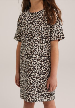 Enfant portant une robe à manches courtes, longueur genoux, avec un motif léopard beige et noir, debout devant un fond uni.