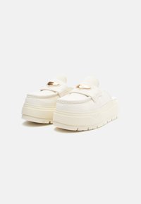 Off-white suedette plattform slip-on loafers med tykke såler og metalliske gullmerkelapper på de øvre remmene, satt mot en hvit bakgrunn.