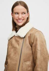 Bruine faux leren jas met een crèmekleurige schapenvachtkraag, ritssluiting en zijzak. Gladde textuur, oversized pasvorm en casual ontwerp.