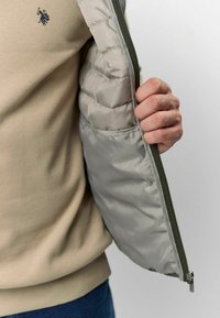 Bež dukserica s tamnim logom, uparena s svijetlosivim pufastim jaknom koja ima glatku teksturu i zatvarač maslinaste boje.