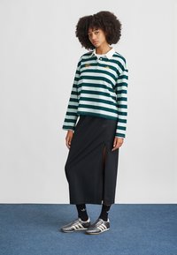 Polo de manga larga a rayas verdes y azules con un collar blanco, combinado con una falda negra con una abertura lateral, zapatillas de fútbol grises y calcetines negros.