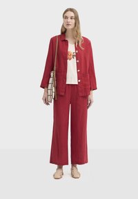 Veste en lin rouge à gros boutons, associée à un pantalon large assorti et un t-shirt blanc à motifs floraux. Des chaussures beiges et un sac à motifs complètent le look.